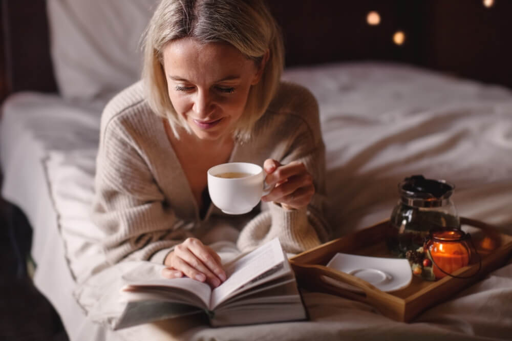 Adult attractive woman is reading book and drinking tea in her bedroom