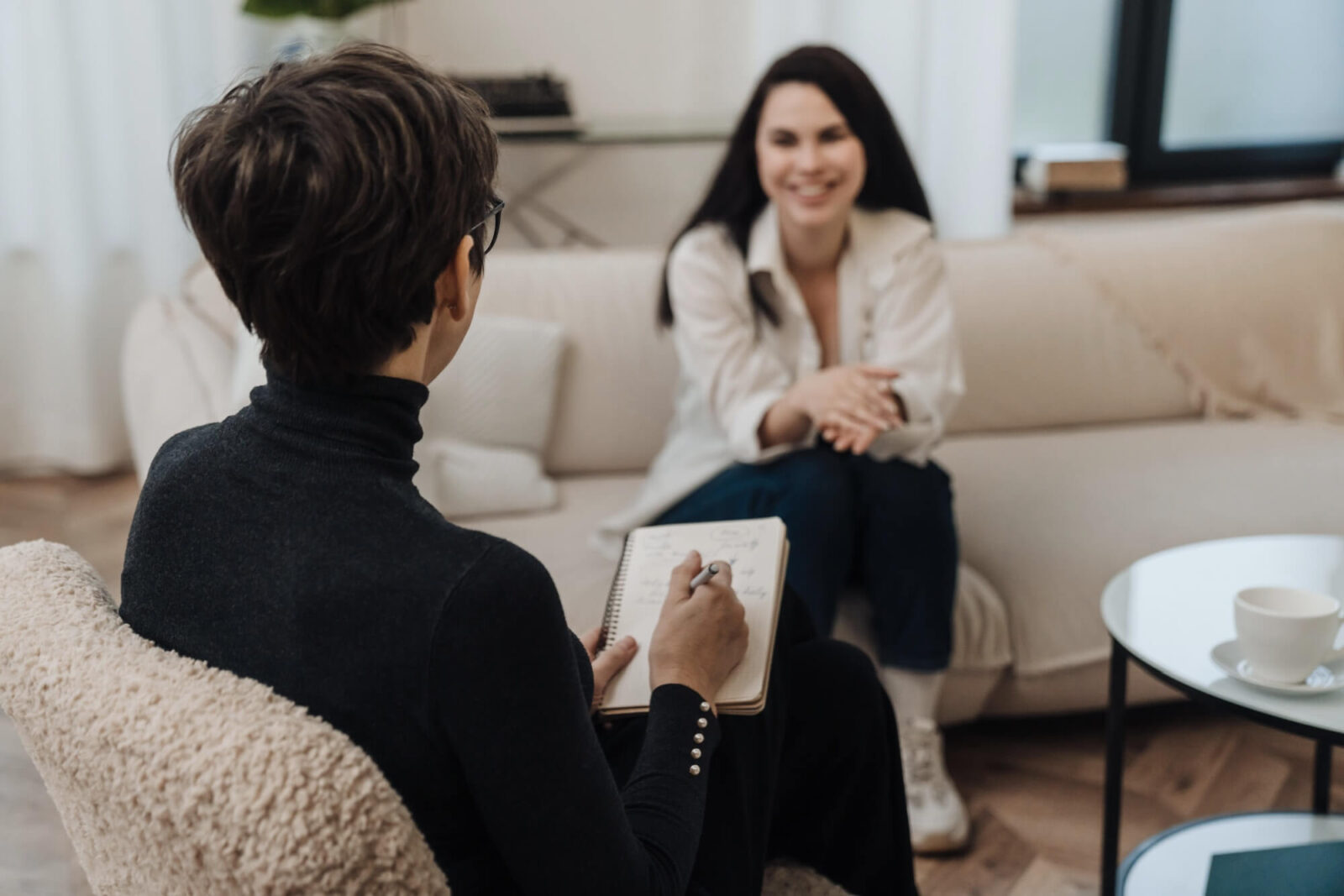 Female psychologist writing down notes during therapy session with smiling young woman