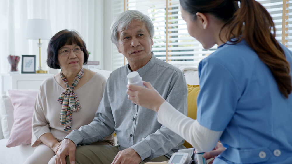Young caregiver in scrubs uniform showing medicine bottle to elderly couple