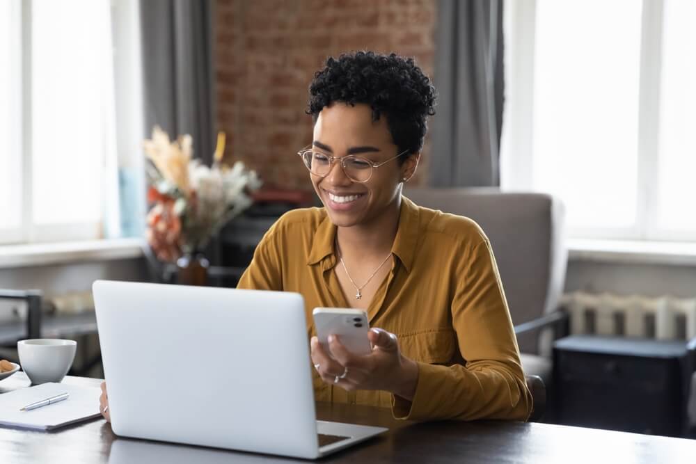 woman sit at workplace desk holds cellphone while using laptop
